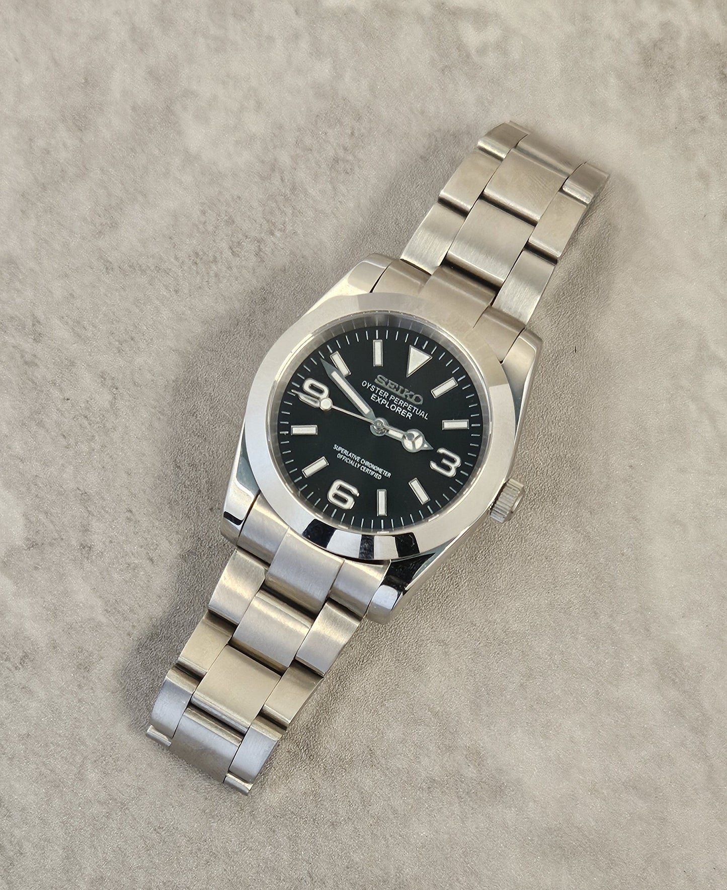 A Seiko Explorer Mod Watch with a black dial and a stainless steel bracelet, featuring a 36mm case with a black explorer style dial, on a textured grey background.