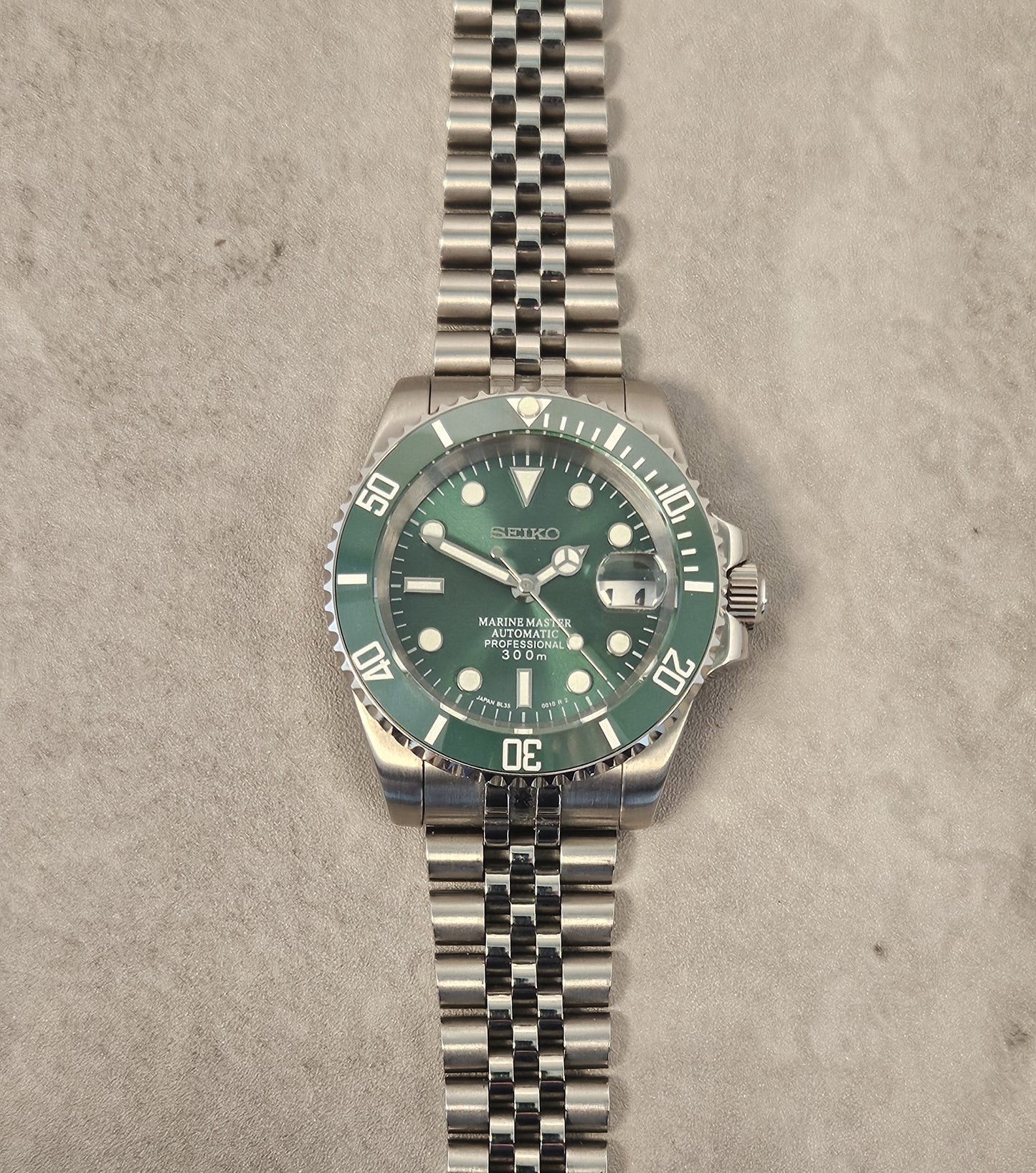 A custom mod Seiko watch with a green dial and green ceramic bezel, a silver stainless steel bracelet, displayed on top of a passport with a pen next to it.