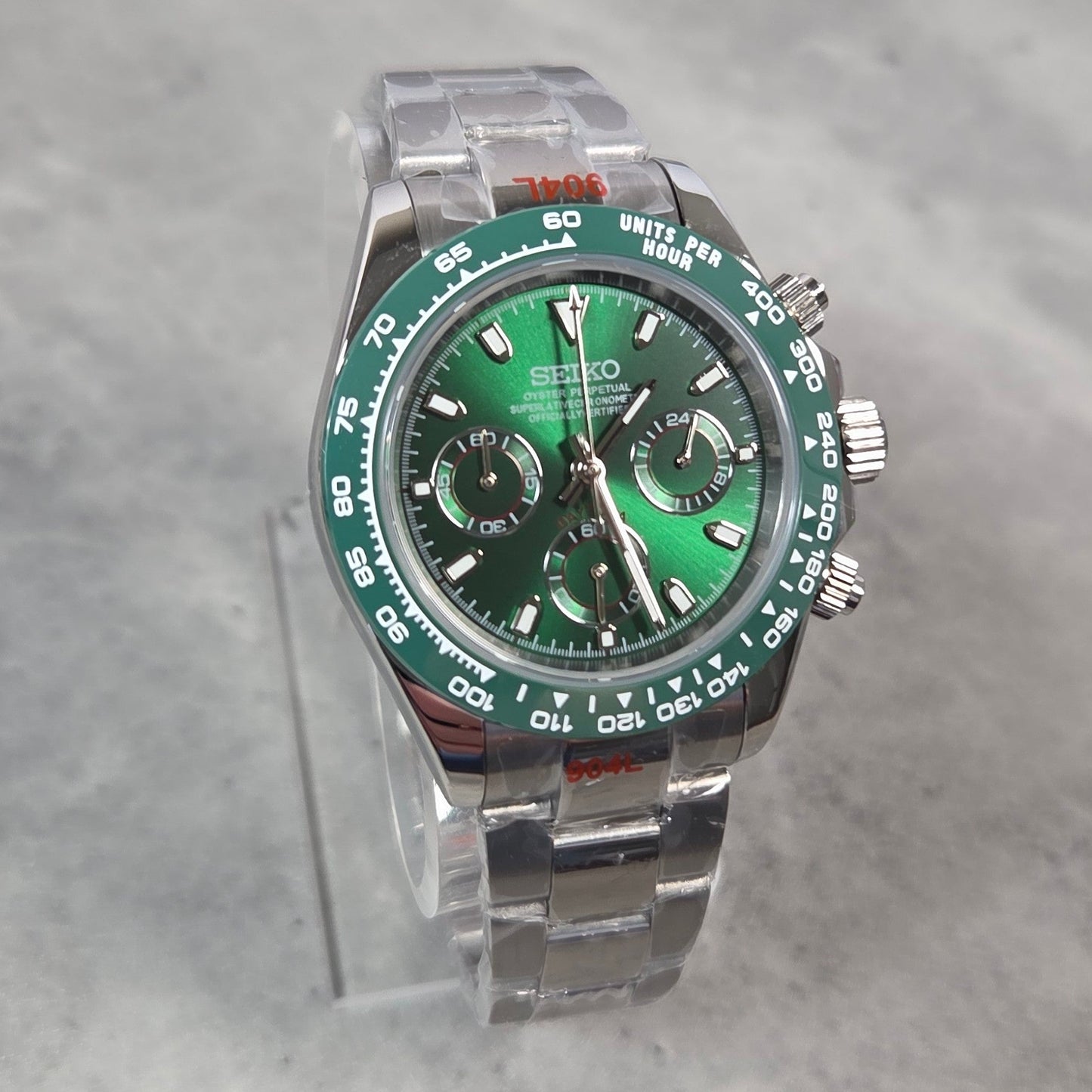 A Seiko stainless steel watch with a green dial and green tachymeter bezel, displayed on a clear acrylic stand against a gray marble background.