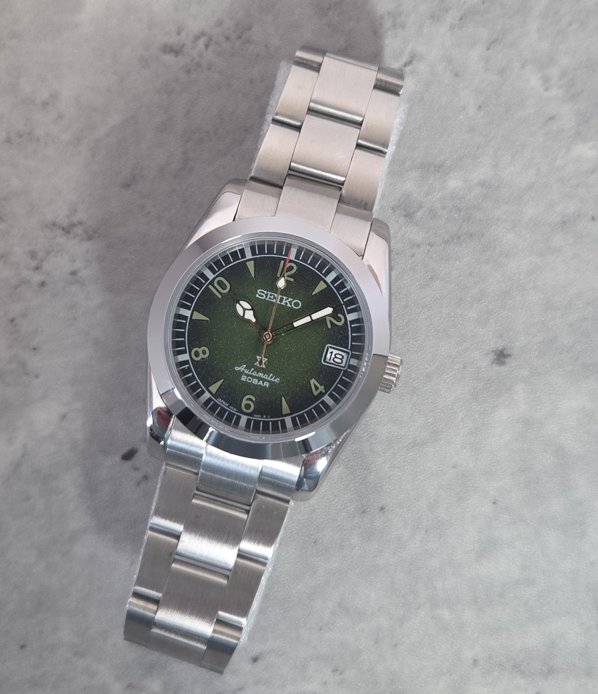 A Seiko Automatic Solar wristwatch with a green dial and a silver stainless steel link bracelet, photographed on a textured gray surface.