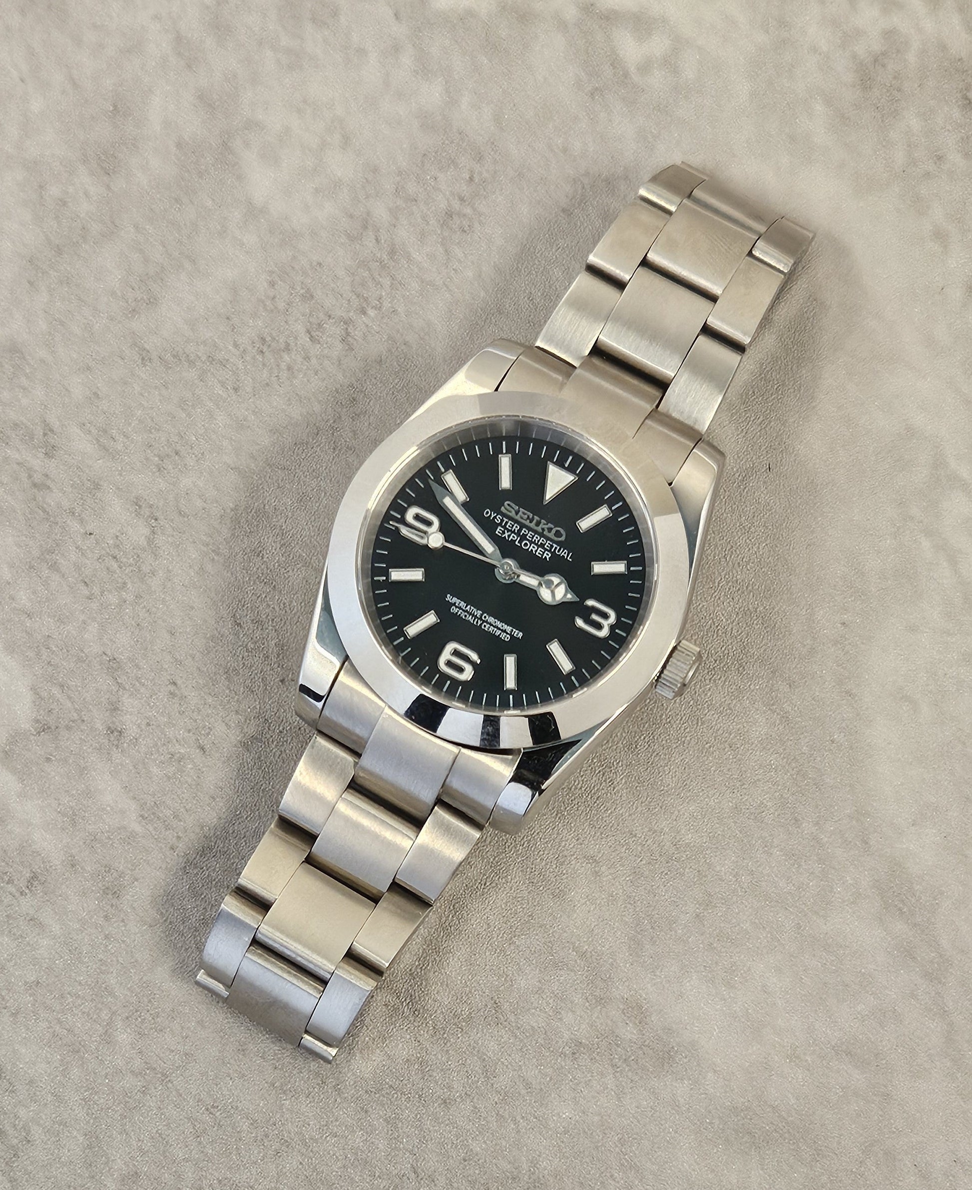 A Seiko Explorer Mod Watch with a black dial and a stainless steel bracelet, featuring a 36mm case with a black explorer style dial, on a textured grey background.