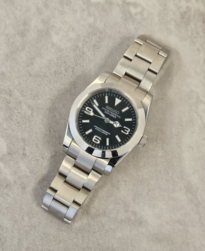 A Seiko Explorer Mod Watch with a black dial and a stainless steel bracelet, featuring a 36mm case with a black explorer style dial, on a textured grey background.