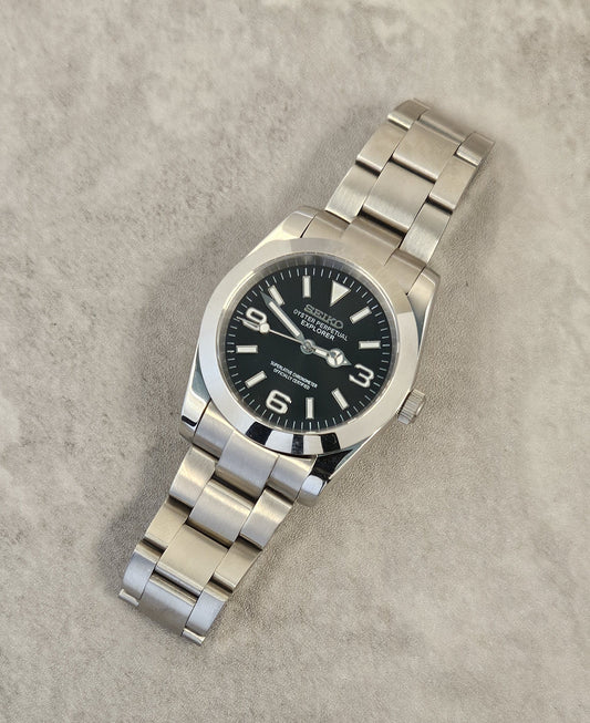 A Seiko Explorer Mod Watch with a black dial and a stainless steel bracelet, featuring a 36mm case with a black explorer style dial, on a textured grey background.