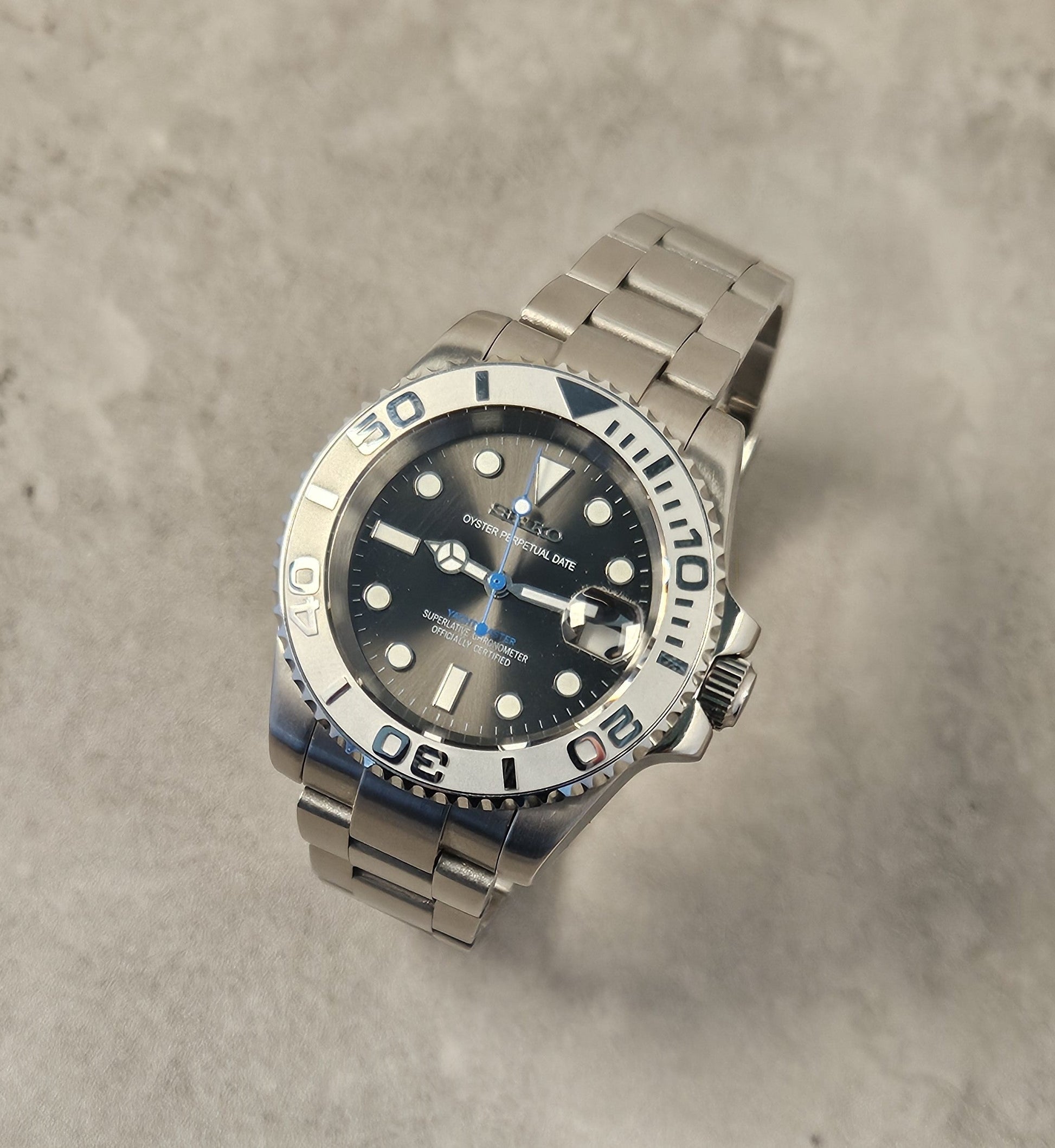 A luxurious silver-toned watch with a grey sunburst dial, luminous hands, a unidirectional rotating bezel, and a stainless steel bracelet on a textured background.