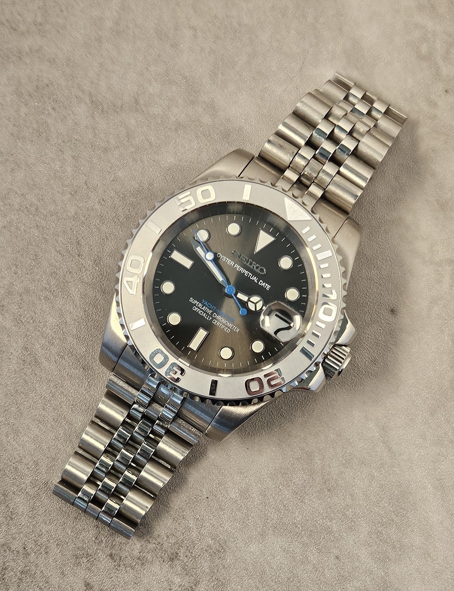 A luxurious silver-toned watch with a grey sunburst dial, luminous hands, a unidirectional rotating bezel, and a stainless steel bracelet on a textured background.