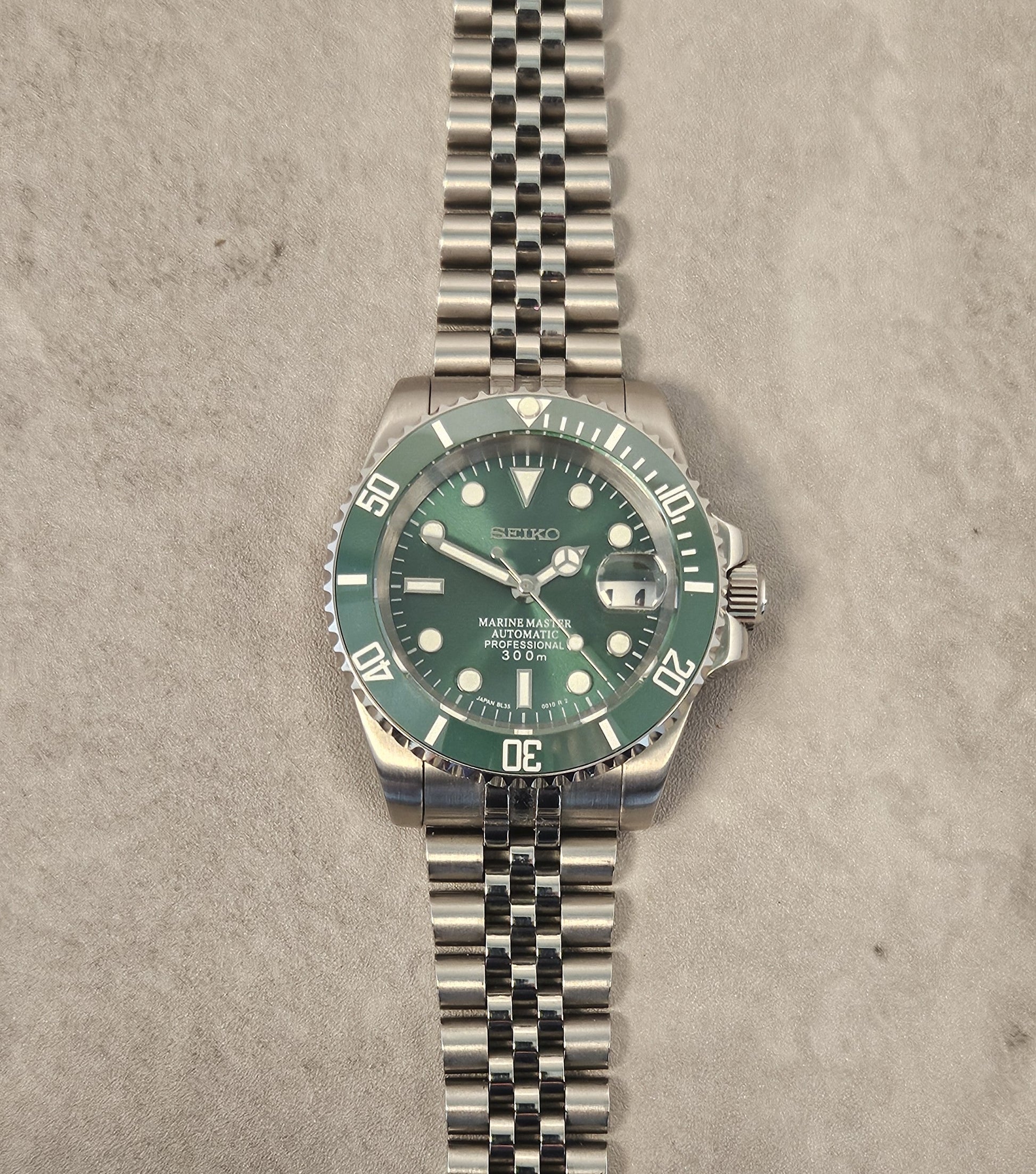 A custom mod Seiko watch with a green dial and green ceramic bezel, a silver stainless steel bracelet, displayed on top of a passport with a pen next to it.