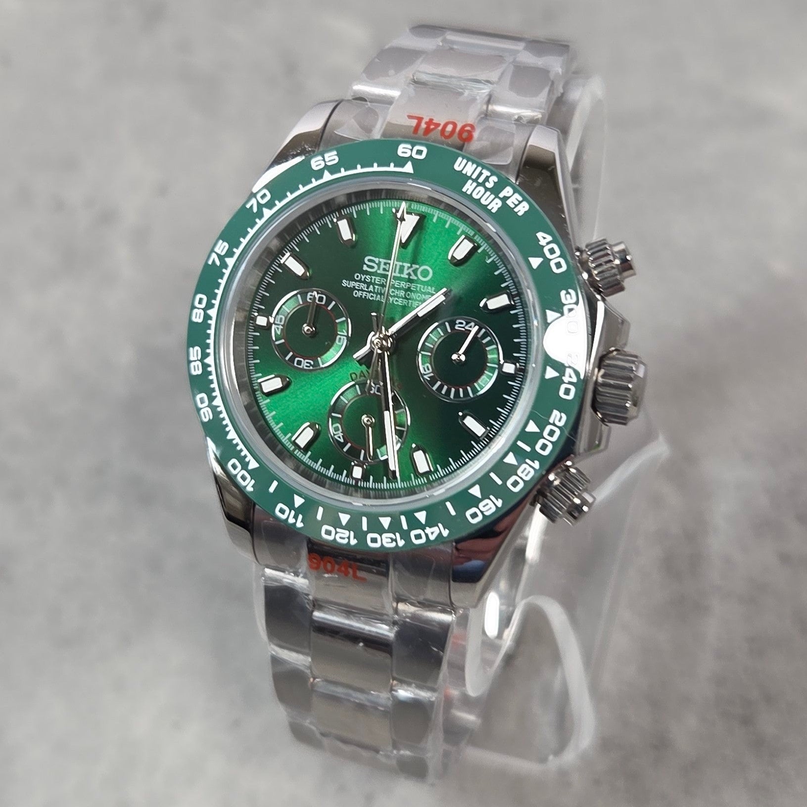 A Seiko stainless steel chronograph watch with a green dial and green bezel, displayed on a clear stand against a light gray textured background.