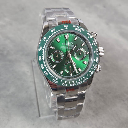 A Seiko stainless steel watch with a green dial and green tachymeter bezel, displayed on a clear acrylic stand against a gray marble background.