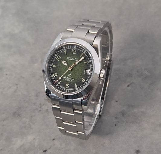 A Seiko stainless steel wristwatch with a green dial and silver link bracelet, photographed on a textured gray concrete surface.