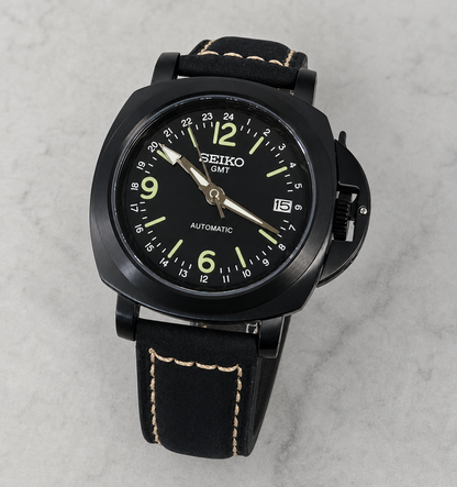 A Seiko GMT automatic watch with a black case, black dial, and black leather strap with light tan contrast stitching, photographed on a light gray marble surface.