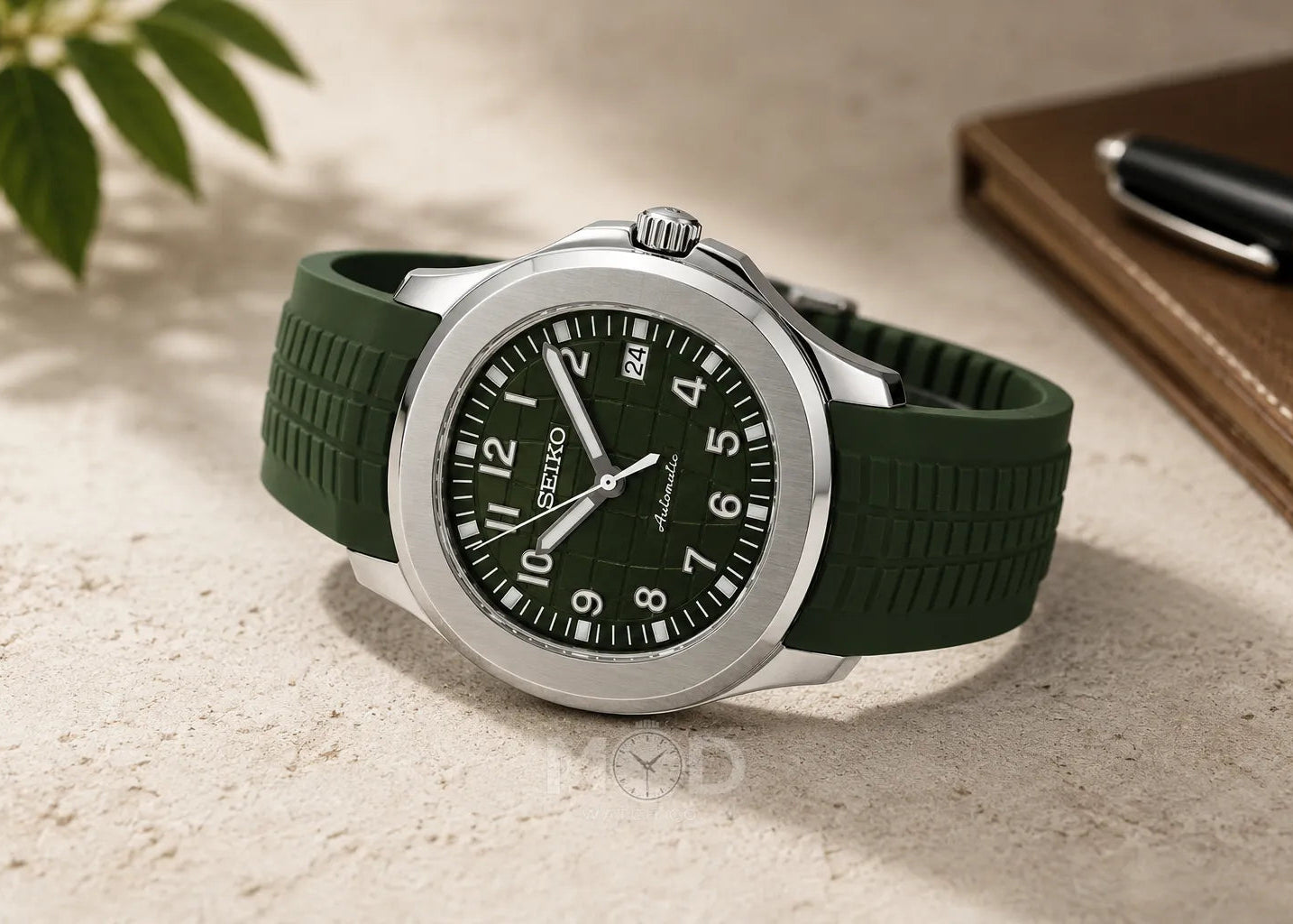 Green wristwatch with a black dial on a light surface, next to a pen and notebook.