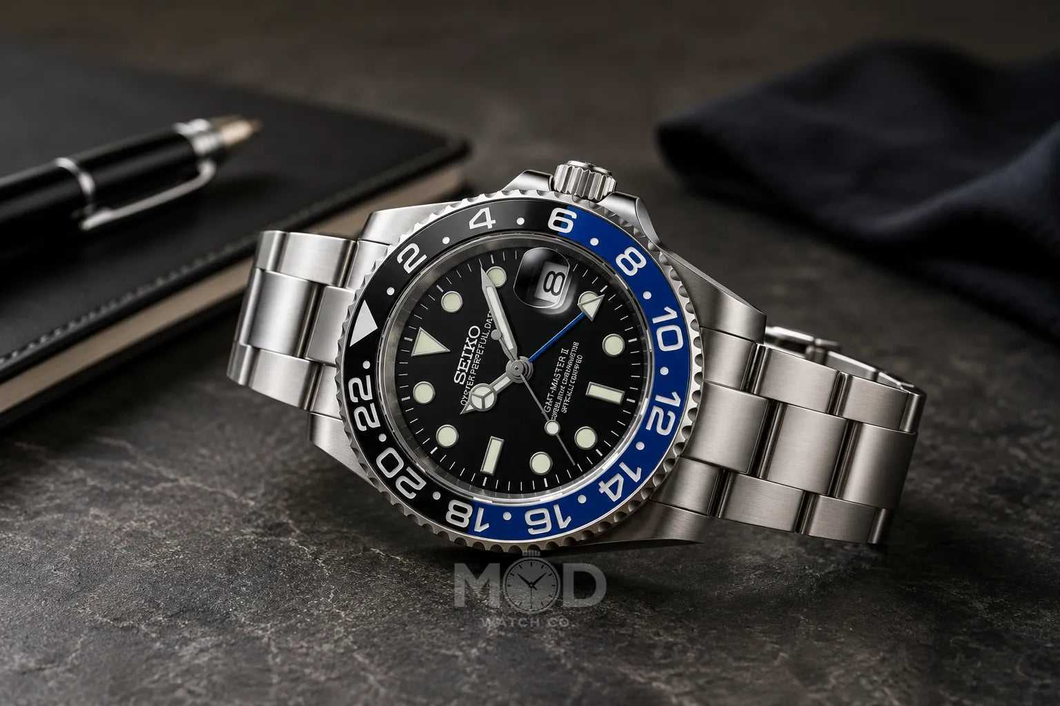 Silver watch with a black and blue face on a dark surface, with a pen and notebook in the background.