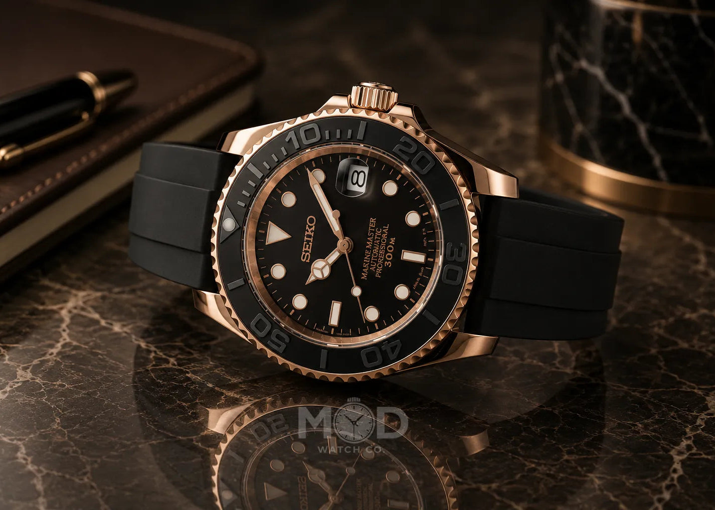 Black and rose gold watch on a reflective surface with a pen and notebook in the background