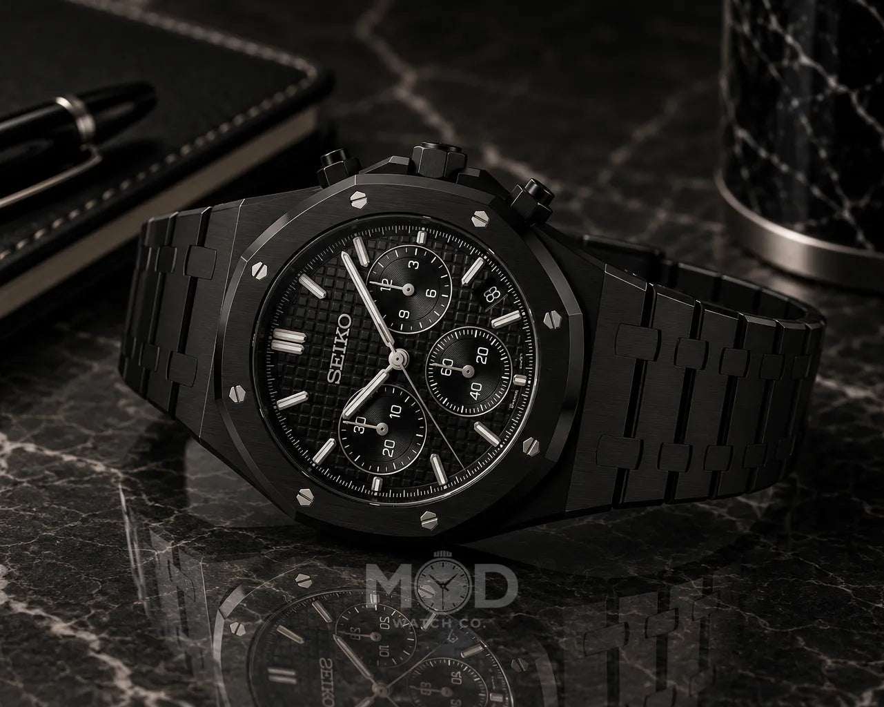 Black Seiko watch on a dark marble surface with a pen and container.
