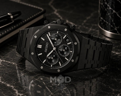 Black Seiko watch on a dark marble surface with a pen and container.