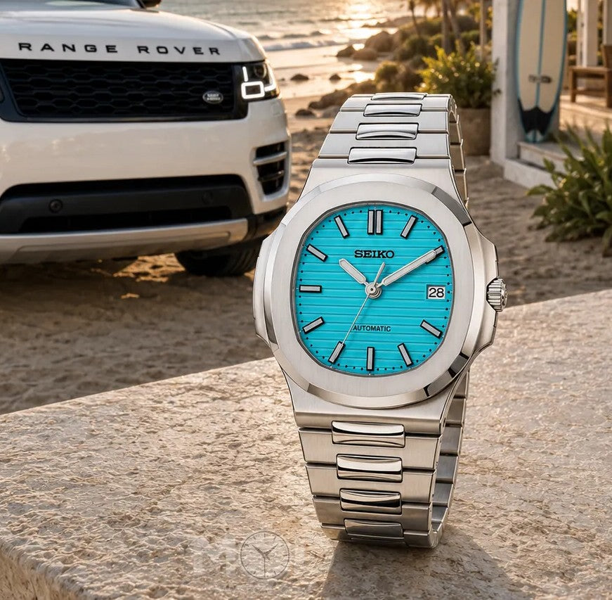 Silver Seiko watch with blue face on a stone surface, Range Rover in the background.