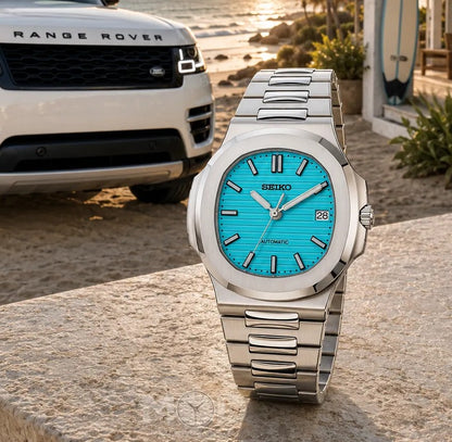Silver Seiko watch with blue face on a stone surface, Range Rover in the background.