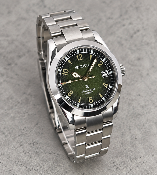 A Seiko Automatic 100m stainless steel watch with a dark olive green dial and matching green accents, displayed on a textured gray surface.