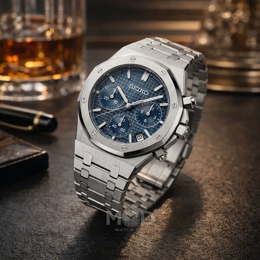 Silver Seiko watch with blue face on a dark surface with a glass of whiskey in the background.