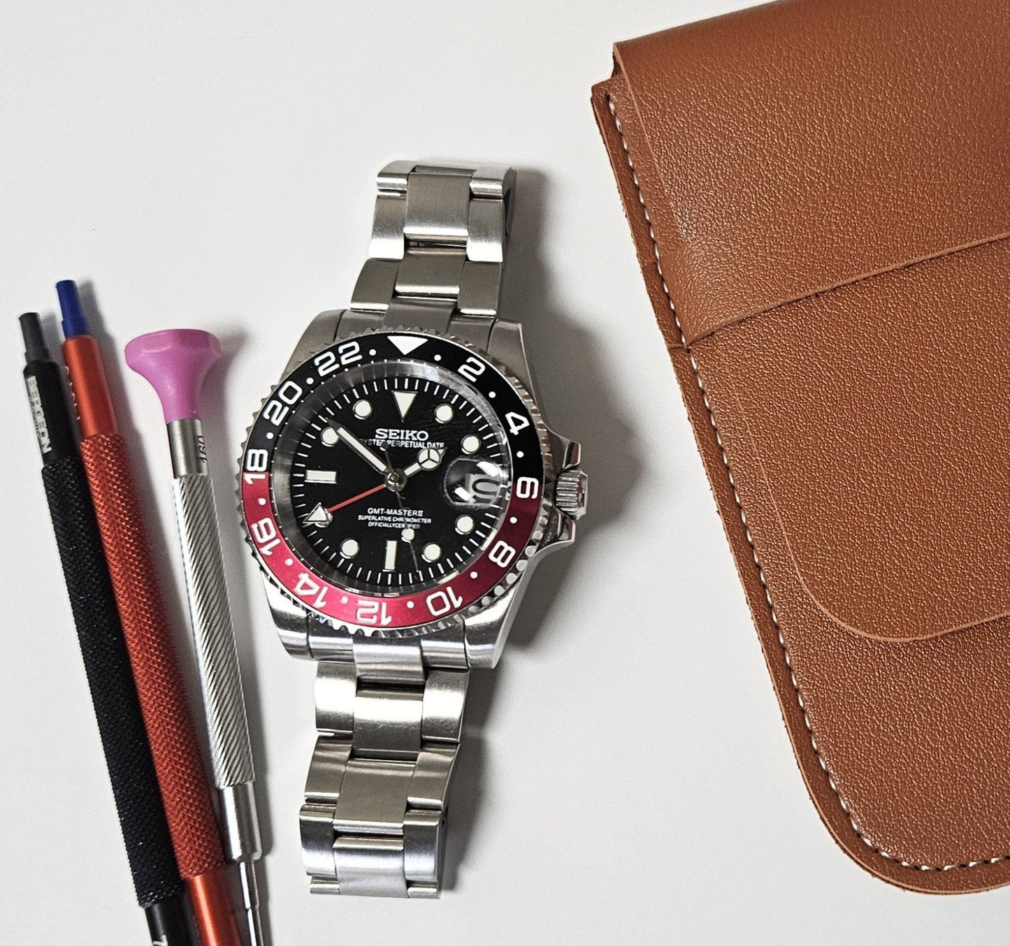 A GMT Master custom mod watch with a black dial, black and red bezel, and silver bracelet, displayed alongside a brown leather case, a cleaning brush, and a watch pen.