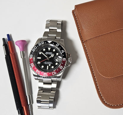 A GMT Master custom mod watch with a black dial, black and red bezel, and silver bracelet, displayed alongside a brown leather case, a cleaning brush, and a watch pen.