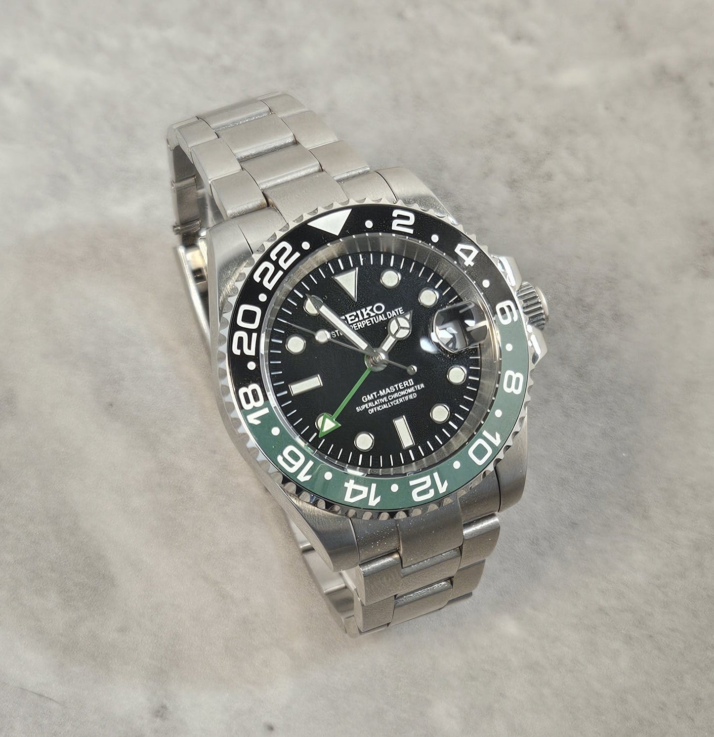A GMT Master Spite Custom Mod Watch with a black dial, black and green bezel, and a stainless steel bracelet laid out next to a cup and pens.