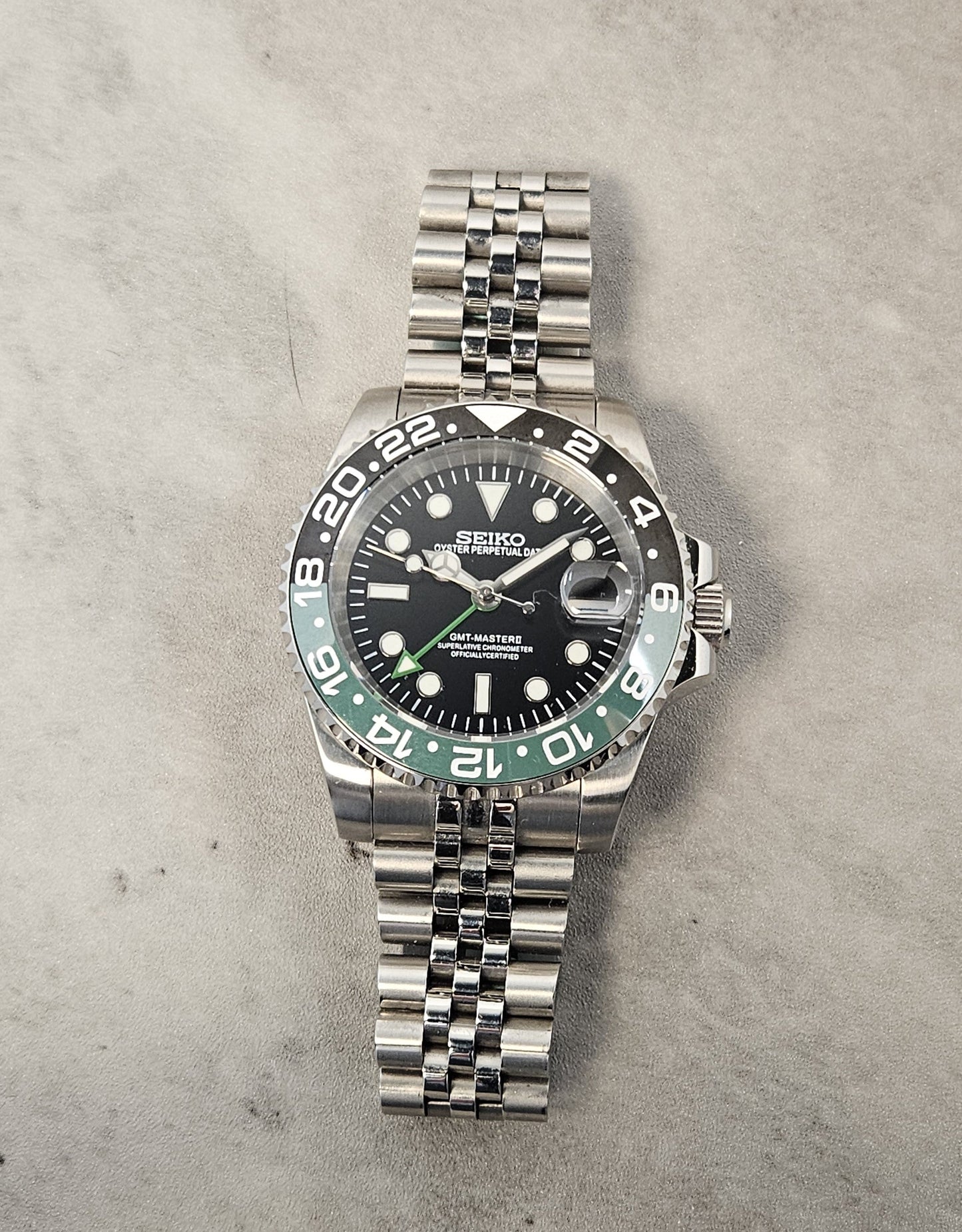 A GMT Master Spite Custom Mod Watch with a black dial, black and green bezel, and a stainless steel bracelet laid out next to a cup and pens.